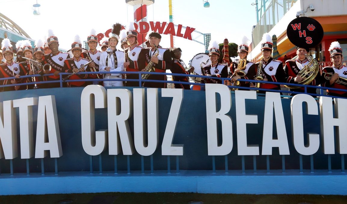 Woodside High marching band in orange and black uniforms performing together atop the Santa Cruz Beach Boardwalk sign
